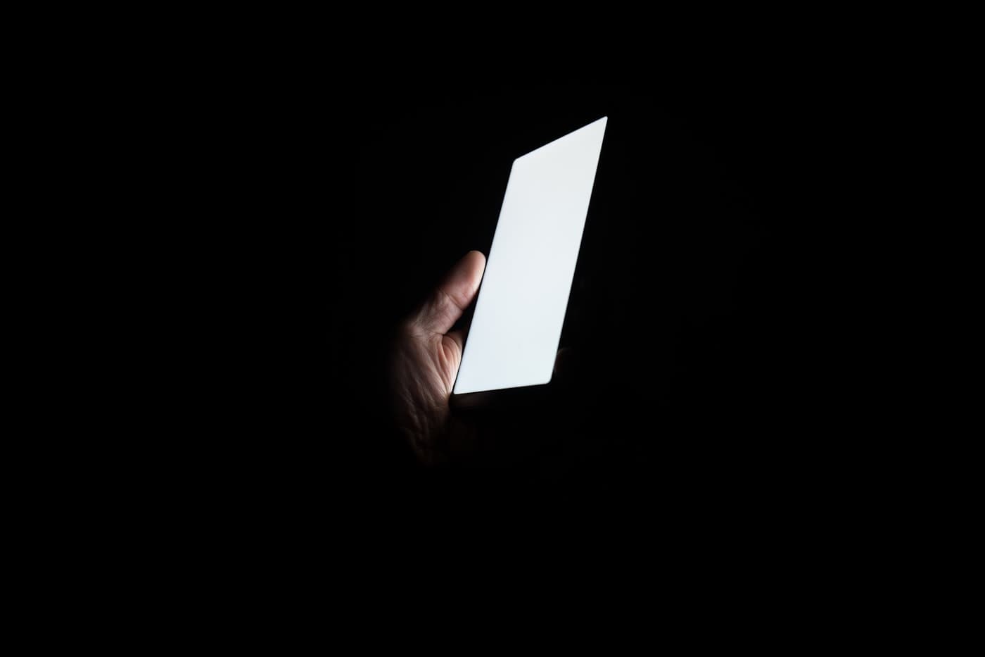 A hand holding a phone with a blank glowing screen in a completely dark room.